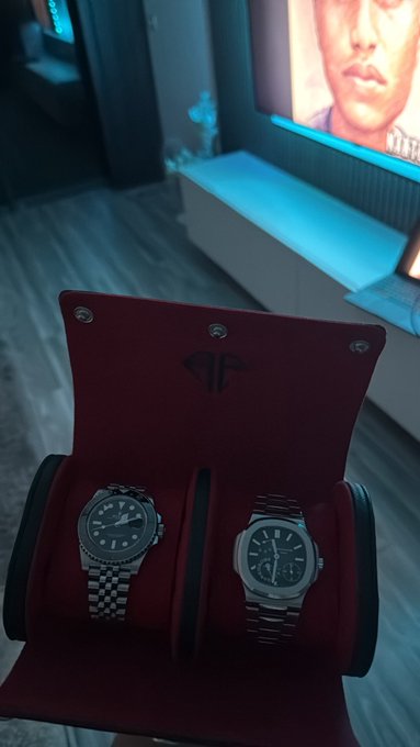 Two luxury watches sit side by side in an open red velvet display box in a room lit with blue ambient light.