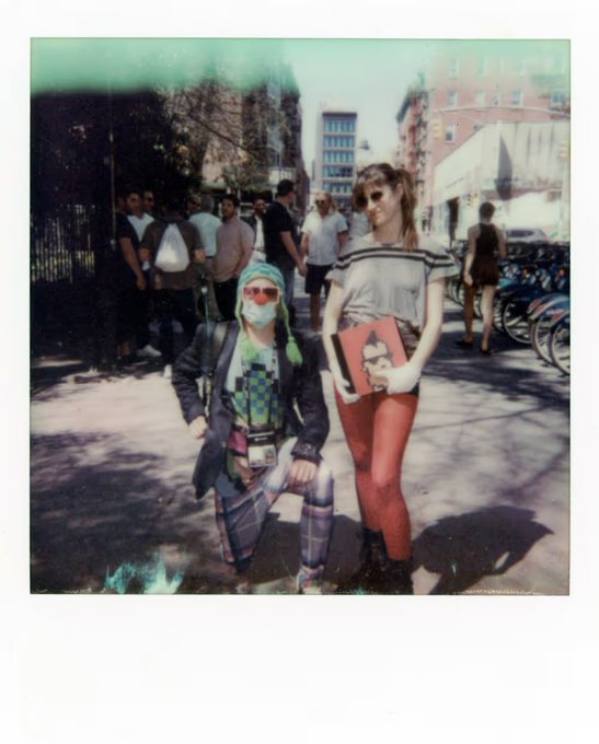 A Polaroid-style photo showing two people walking down a city street — one wearing a surgical mask, sunglasses, and a backpack, the other with long brown hair holding a notebook and wearing red leggings.