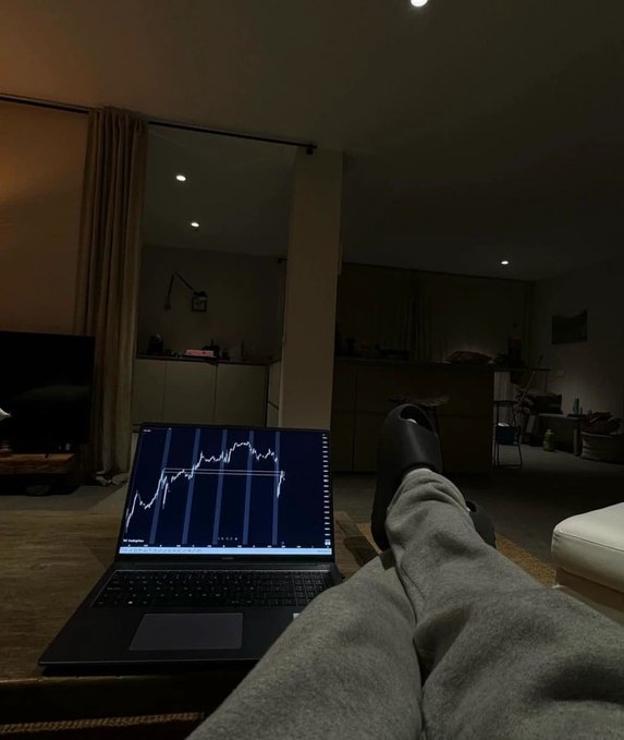 A person relaxes in a dimly lit living room with their feet propped up, displaying a laptop screen showing a candlestick trading chart that appears to spike upward, visually reinforcing their post about stealth-launching a Solana memecoin called "cupracall" and giving away 10,000,000 tokens to the first 999 retweeters.