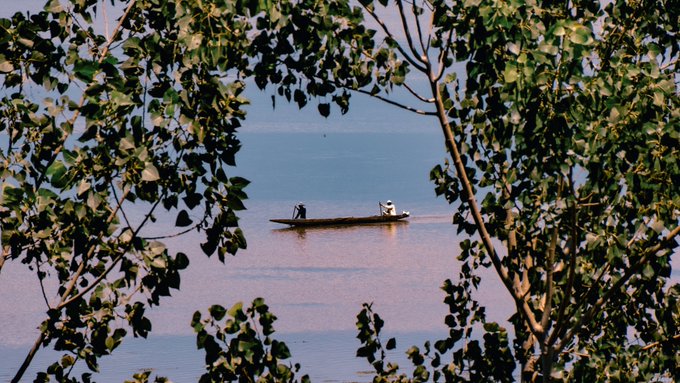 Two people in a small wooden boat floating on a calm river, viewed through a natural frame of green leaves and tree branches.