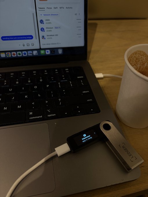 A Ledger hardware wallet connected via USB to a MacBook displaying a wallet interface, sitting next to a paper coffee cup.
