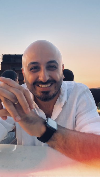 A selfie of a bald man with a dark beard, wearing a white shirt and a black smartwatch, smiling at the camera against a sunset sky.