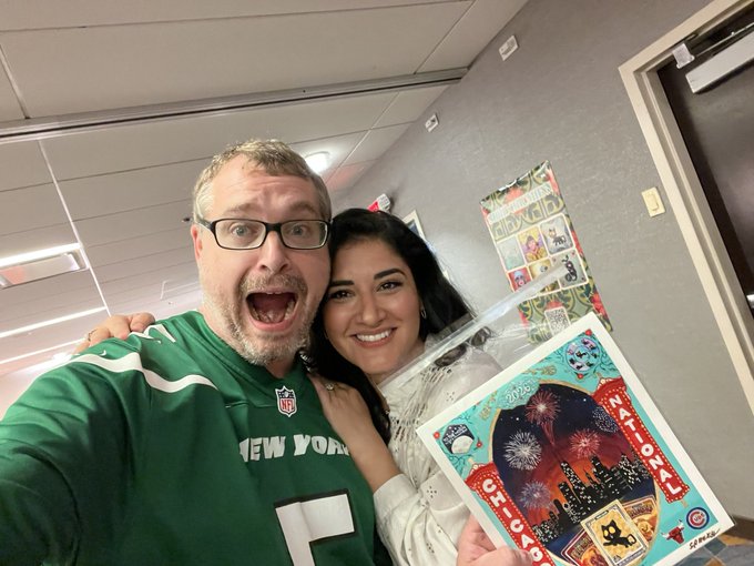 A man wearing a green New York Jets jersey and glasses smiles with a woman holding a wrapped Christmas-themed gift and a card dated 12-10-2022.