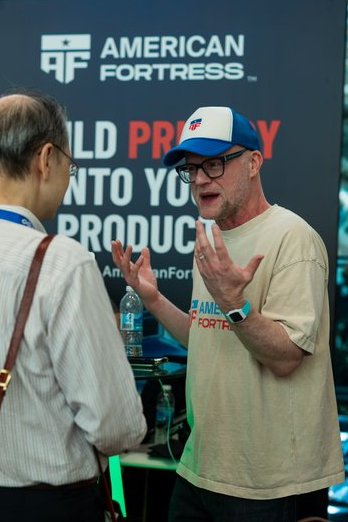 A man in a tan "American Fortress" t-shirt and red, white, and blue cap speaking animatedly at what appears to be an expo booth.
