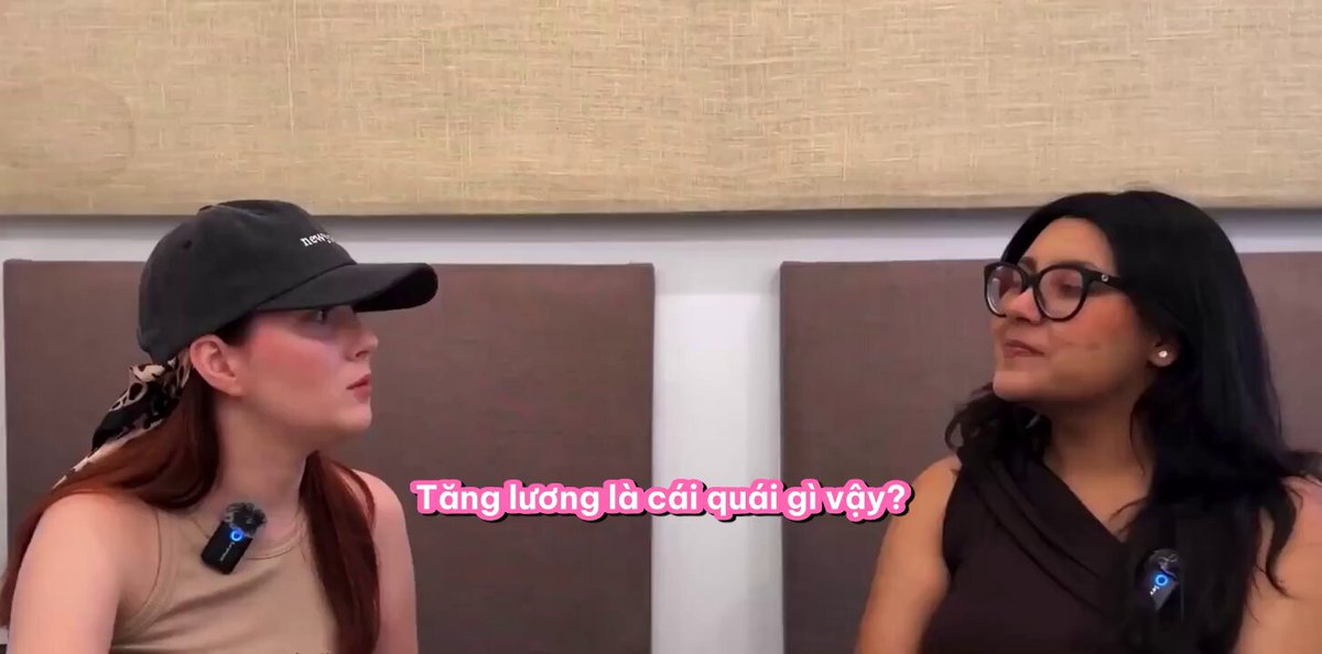 The image shows two women sitting next to each other, engaged in a conversation. They are both wearing hats and glasses, which adds an interesting visual element to the scene. The background of the image features a brown wall with text written on it that reads "TANG KýING LÀ NƠI NHỮNG SỰ THẬT TRẦN TRỤI NHẤT Hôm qua Kim ngồi xem chart, thấy 𝐁𝐓𝐂 65,752 USD lướt nhẹ âm 𝟎&𝐆 22 là mức Extreme Fear khiến cả group Telegram im phâng phắc.