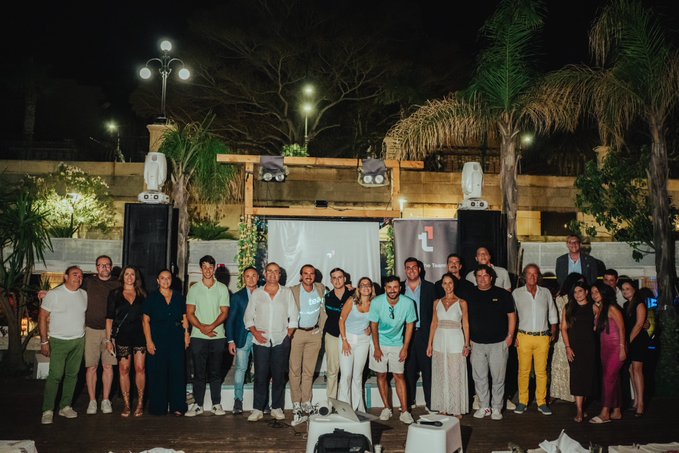 A group photo of roughly twenty people standing outdoors at night at an event, with palm trees, lights, and equipment visible in the background.