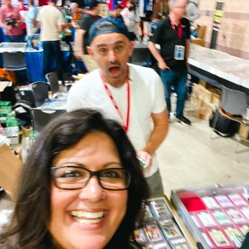 A selfie of a smiling woman wearing glasses taking a photo with a man behind her who has an excited expression, appearing to be at a trading card or collectibles convention.