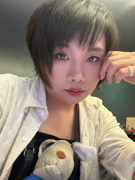 A selfie of a young woman with short dark hair wearing a light-colored button-up shirt, holding a plush toy in a carrier.