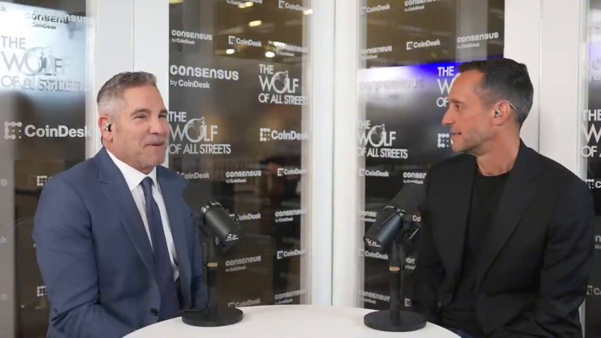 The image shows two men sitting at a table, each wearing a suit and tie. They are engaged in an interview or discussion with a microphone set up between them. The background features a glass wall that reflects their surroundings, creating a sense of depth and context for the scene.