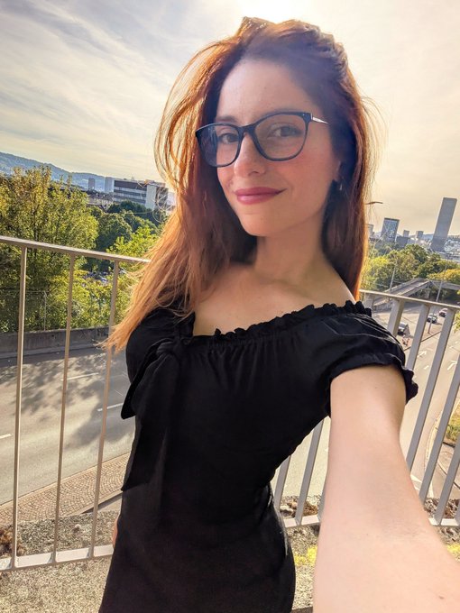 A young woman with reddish-brown hair wearing black cat-eye glasses and a black off-shoulder top takes a selfie outdoors on a balcony, with trees and city buildings visible in the background.
