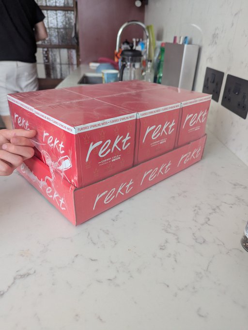 A hand unwrapping shrink wrap from a stack of pink 'rekt' cherry cola 12-pack boxes on a white marble countertop.
