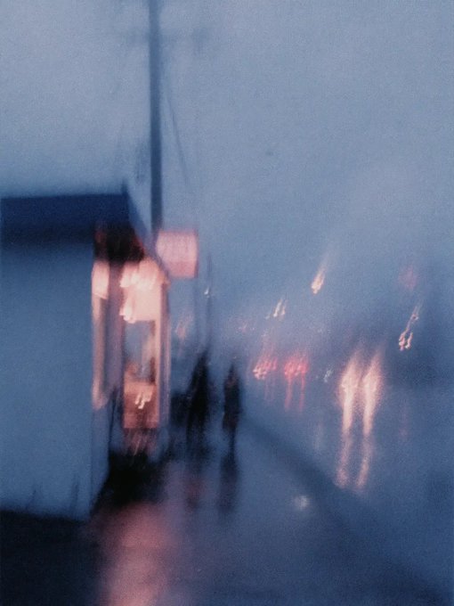 A blurred, atmospheric photograph shows two figures walking down a foggy street with glowing lights reflecting on the wet pavement.