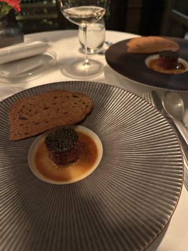 A fine dining presentation featuring what appears to be caviar atop a small portion of food on a textured glass plate, with bread and a wine glass in the background.