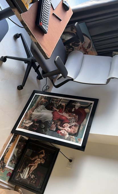 An overhead view of a home office with a desk holding a keyboard, an office chair, and framed classical paintings leaning against the wall, including one resembling Botticelli's work.