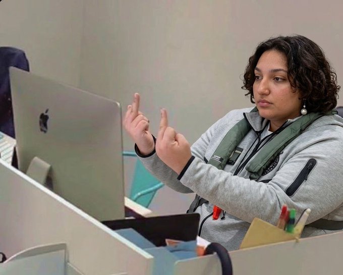 A woman with dark curly hair sits at a desk gesturing with both hands, working in front of an Apple iMac monitor in what appears to be an office or collaborative workspace.