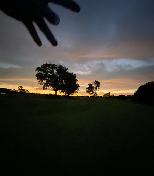 A dark silhouette of a hand reaching into the frame against a dramatic sunset sky over a golf course with trees and a yellow flag.