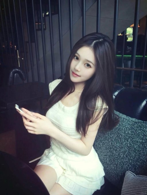 A young woman with long dark hair wearing a white sleeveless top and skirt sits on a dark sofa holding a smartphone, smiling at the camera in what appears to be a cafe or lounge setting.