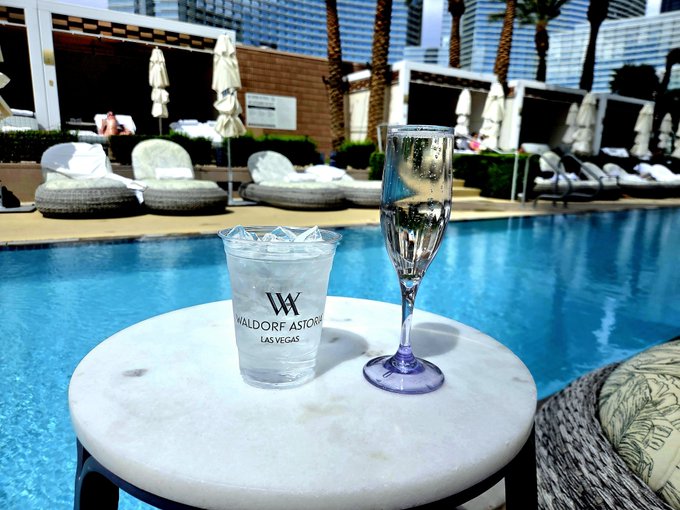 A champagne flute and a "Waldorf Astoria Las Vegas" branded tumbler with ice sit on a marble-topped table overlooking a luxurious pool area with cabanas and palm trees, capturing the post's celebration of "PROFITS last cycle" and the user's claimed exclusive access to the Waldorf Astoria pool.
