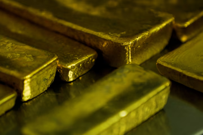 A close-up photograph shows several stacked gold bars with a reflective surface and some water droplets on them.