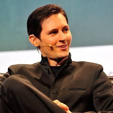The image features a man wearing a suit, sitting down with his legs crossed. He has short hair and is smiling at the camera while looking off into the distance. The caption of the image reads "BREAKING: TON'S native currency is rebranding to Gram TON remains the name of the blockchain this is step 4/7 to Make $TON Great Again Durov's cooking something". This post seems to be discussing a recent development in the cryptocurrency world, specifically related to the Dollar token. The image captures the man's facial expression and body language as he engages with the content displayed on the screen.