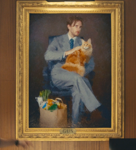 An oil painting in a gold frame titled 'GUS' depicting a man in a blue suit holding an orange cat, with a grocery bag of vegetables at his feet.