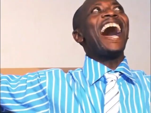 A man wearing a blue and white striped shirt with a striped tie is laughing heartily, looking upwards with his mouth wide open.