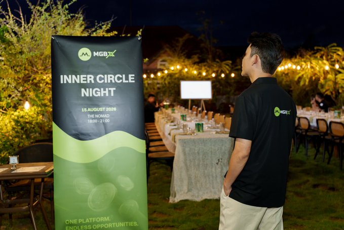 A man wearing an MGBX-branded black polo stands near a banner reading "MGBX INNER CIRCLE NIGHT, 15 AUGUST 2026, THE NOMAD, 18:00 - 21:00" with the tagline "ONE PLATFORM. ENDLESS OPPORTUNITIES," overlooking an outdoor dinner setup adorned with string lights, capturing a moment from the MGBX Vietnam event.