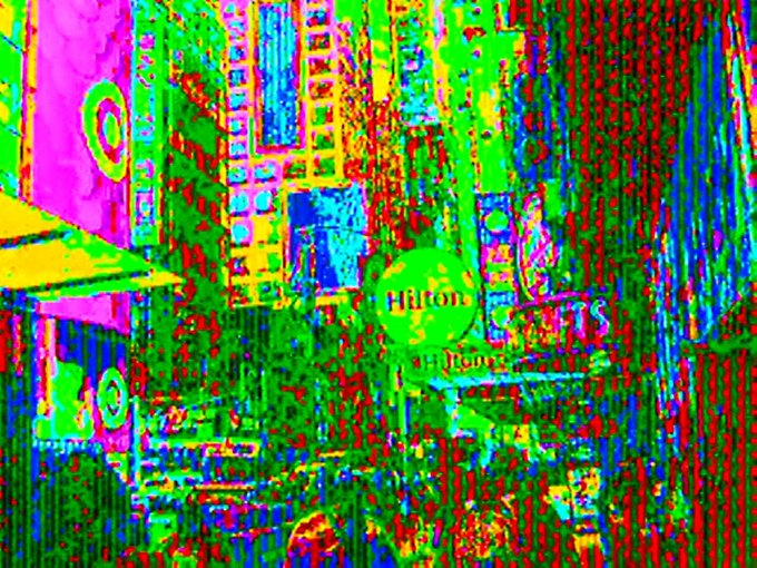 A heavily distorted, glitchy image of Times Square with a 'Hilton Garden Inn' sign visible amid colorful pixelated noise.