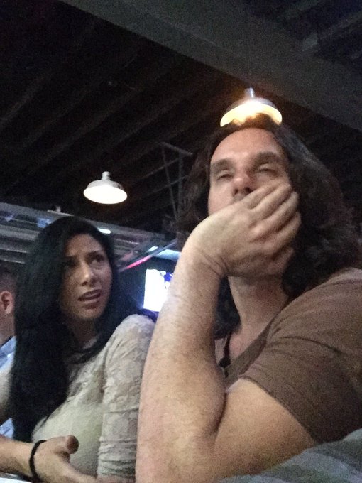 A man in a brown shirt covers his face with his hand in apparent exasperation or frustration, sitting next to a woman with long dark hair who wears a concerned expression at what appears to be a restaurant with hanging pendant lights. The image humorously illustrates the post's punchline, with the man's face-palming gesture perfectly embodying the girlfriend's frustration at her boyfriend's stubbornly bullish stance after selling his crypto early.