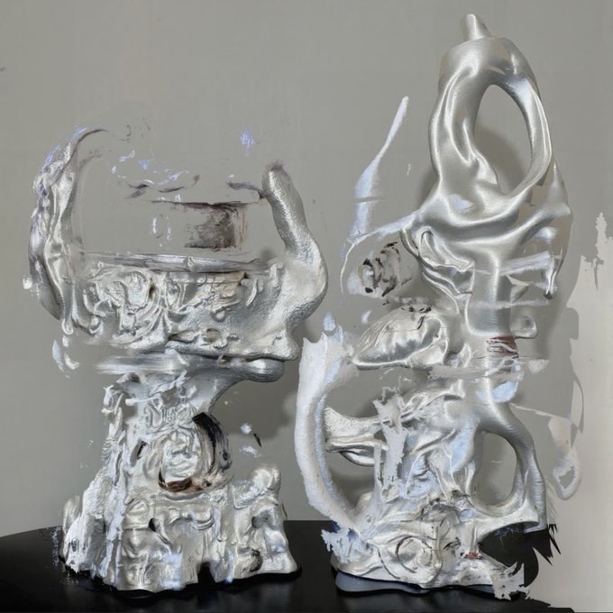 A photograph of two abstract, highly reflective silver sculptures sitting on a dark surface against a plain background, showcasing distorted, liquid-like forms.