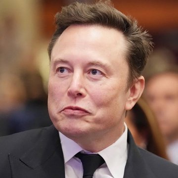A close-up photograph of Elon Musk wearing a dark suit and tie, looking off-camera with a serious expression.