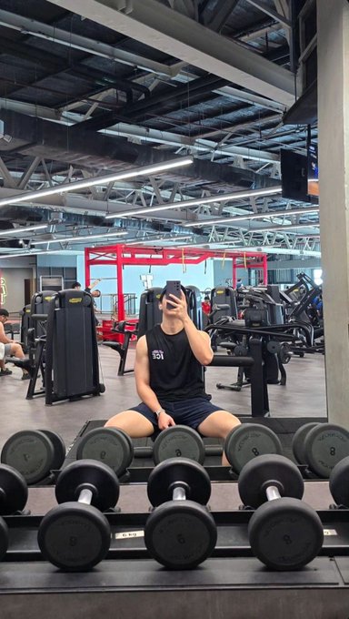 A man takes a mirror selfie in a gym, wearing a black tank top and shorts, kneeling behind a rack of dumbbells.
