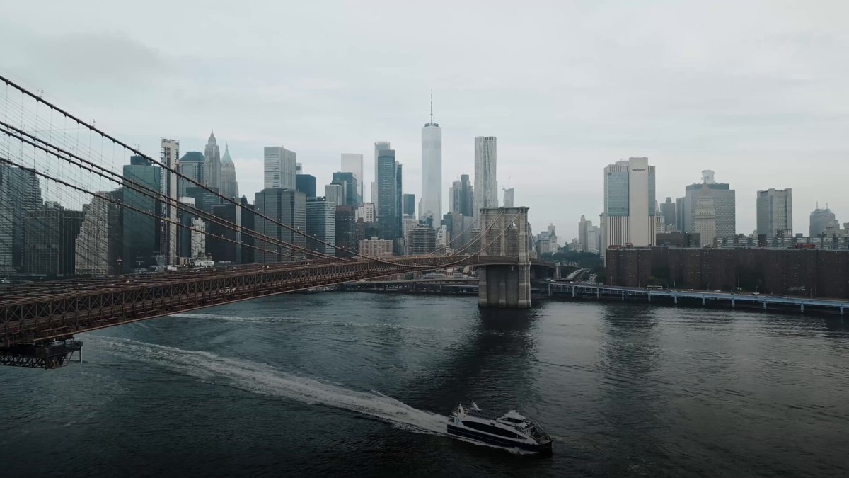 The image shows a city skyline with a large bridge spanning across a river, connecting two parts of the city. A boat can be seen traveling down the river below the bridge. The sky above appears cloudy, creating an overcast atmosphere. In the background, there are tall buildings that make up the cityscape.