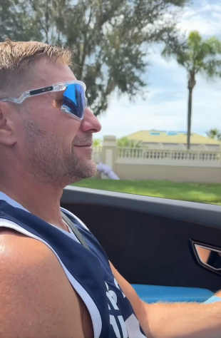 Side profile of a man with blonde hair wearing white Oakley sunglasses and a blue tank top, driving a car with palm trees visible outside.
