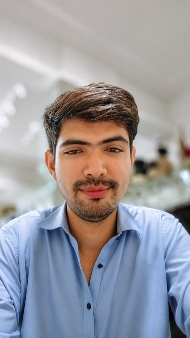 A portrait of a young man with short brown hair, a light beard, wearing a light blue button-up shirt in a well-lit indoor setting.