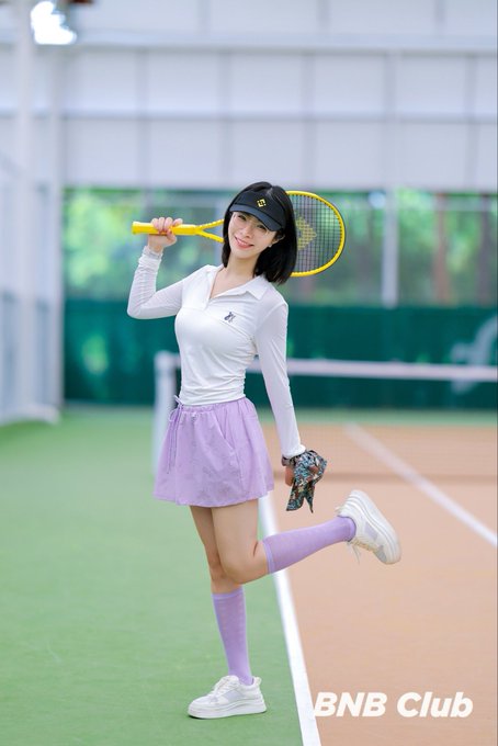 A woman in a white tennis top and lavender skirt posing on a tennis court with a yellow racket, branded with 'BNB Club' watermark.