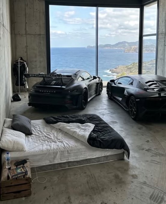 A bedroom with a low mattress on the floor sits directly inside a concrete-walled garage housing two black Porsche sports cars (one a 911 GT3 RS with "PORSCHE" badging), with floor-to-ceiling windows offering a panoramic ocean view—capturing the ultimate luxury of waking up steps from your supercar collection with the sea as your backdrop.