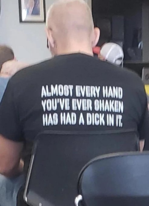 A bald man viewed from behind wears a black t-shirt with the provocative text: 'ALMOST EVERY HAND YOU'VE EVER SHAKEN HAS HAD A DICK IN IT.'