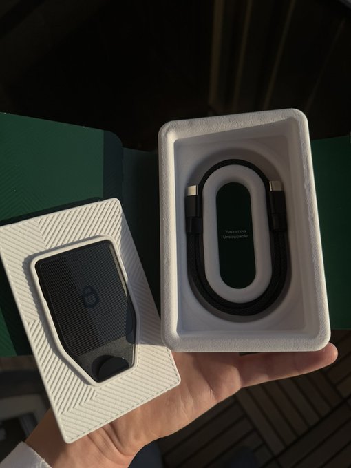 A hand holds an open green Apple product box containing a black Apple Wallet/Card Holder and a USB-C charging cable.