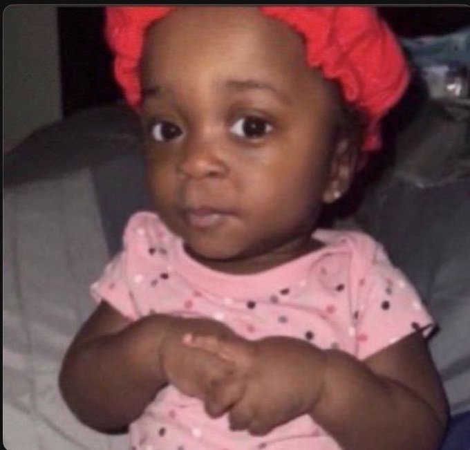 A close-up portrait of a young child with a large red bow in her hair, wearing a pink polka-dot shirt.