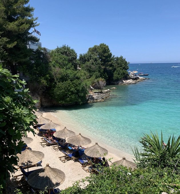 A scenic photograph of a Mediterranean-style cove with crystal-clear turquoise water, a small sandy beach lined with thatched umbrellas and sun loungers, surrounded by lush green trees and rocky cliffs under a clear blue sky.