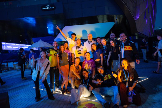 A group of friends pose together at a Solana event, with one person flashing a peace sign and another wearing a race bib reading "181" while sporting a tie-dye shirt. The "Solana" branding visible in the background ties directly to the post's call to tag friends made at a Solana event, capturing the community spirit of the gathering.