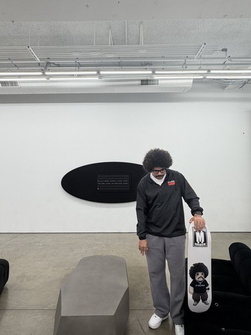 A person with a curly afro and glasses standing in a gallery-like room, holding a skateboard with graphic designs, next to a black oval wall piece.