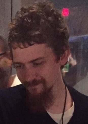 A young man with curly hair, a goatee, and earbuds looks downward with a slight smile.