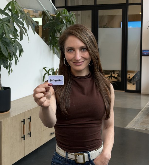A woman in an office holds up a small card labeled “Cognition,” while plants, cabinets, and glass-walled rooms are visible behind her.