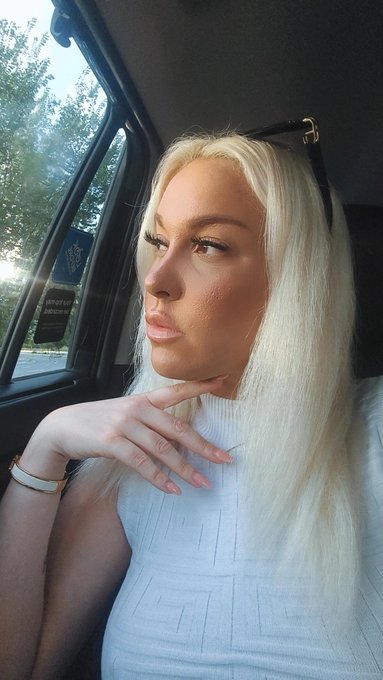 A selfie of a blonde woman with full lips and heavy makeup looking out the window of a car, wearing a white ribbed sleeveless top, with sunglasses resting on her head.