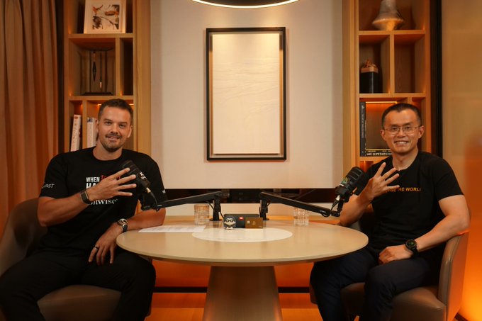Two men sit at a round table with microphones during a podcast recording; the man on the left wears a black "WHEN IN ROME" t-shirt and the man on the right wears a black shirt with orange text. The setup reflects the post's announcement of an upcoming conversation with CZ, Co-Founder of Binance, discussing his journey from building the world's largest crypto exchange to imprisonment and presidential pardon.