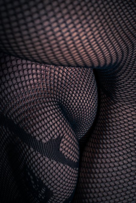 Close-up shot of sheer black fishnet fabric draped in folds, showing the hexagonal mesh pattern in detail. Captured with a Nikon D750 at 50mm (ISO 320, f/6.3, 1/200s), the texture-focused image plays on the saying about leggings telling the truth by revealing the intimate, barely-there nature of the mesh.