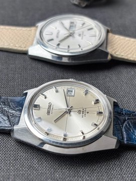 Two Grand Seiko watches displayed on a dark fabric, with the foreground model featuring a silver dial, day-date complications at 3 o'clock, and "Grand Seiko" branding, paired with a navy blue leather strap, while the background watch has a light beige strap.
