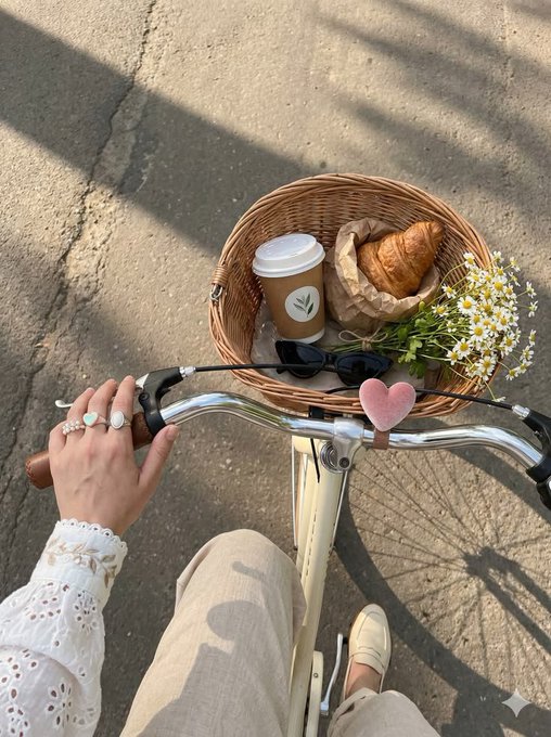 A first-person view of a leisurely bike ride with a wicker basket holding a coffee cup, croissant, and a bouquet of daisies, while the rider's hand—adorned with rings—grips the handlebar next to a small pink heart decoration. The scene captures the peaceful, aesthetic weekend vibe of recharging and enjoying simple pleasures, perfectly complementing the message to take time for self-care.